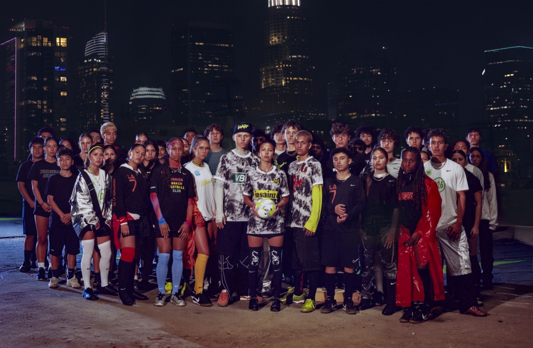 A diverse group of young adults stands together at night in urban sportswear, with city skyscrapers lit up in the background.