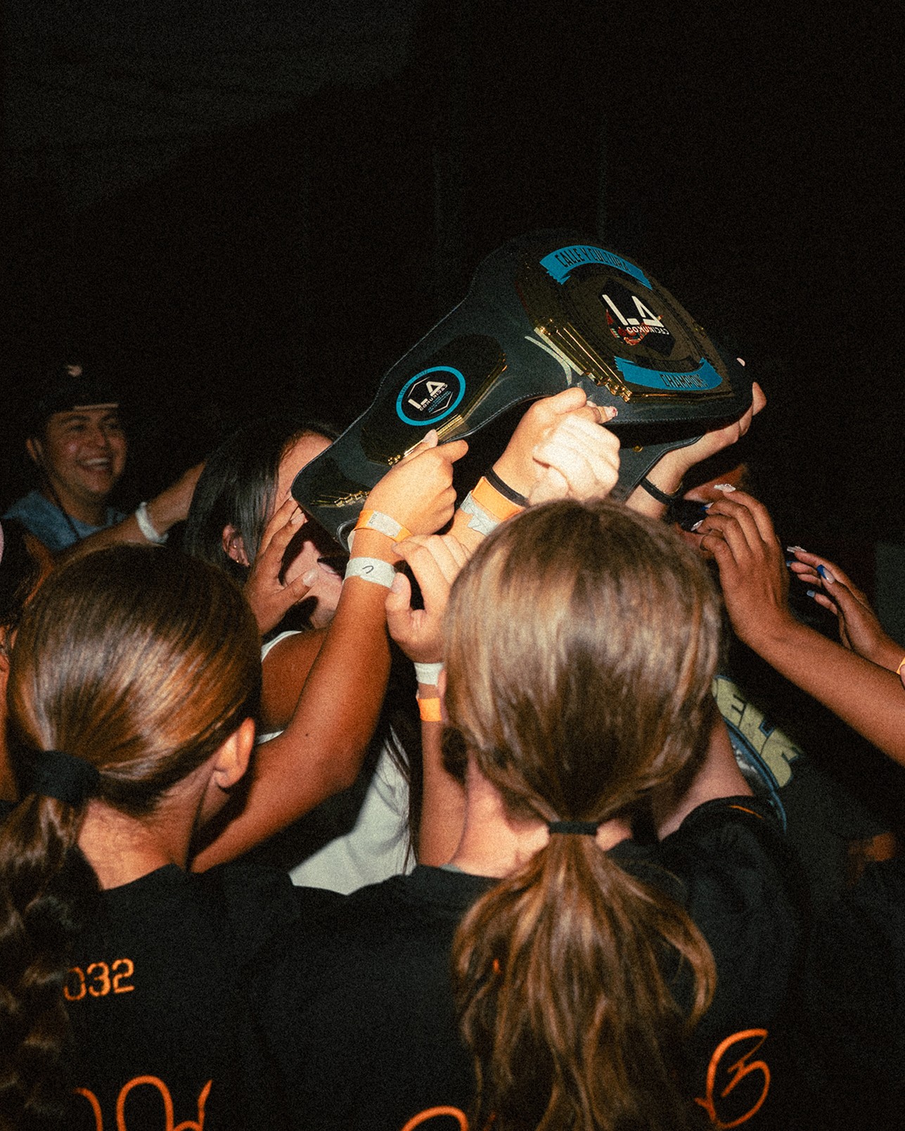 A group of people in black shirts stand close together, holding up a large championship belt in celebration.