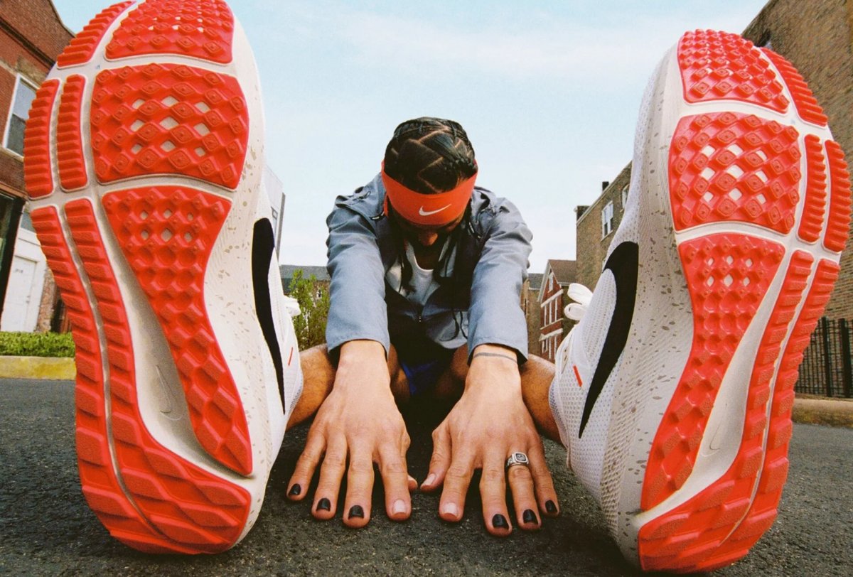 Person stretching on a street, touching their toes, with large focus on the soles of their red and white running shoes; buildings and sky visible in background.