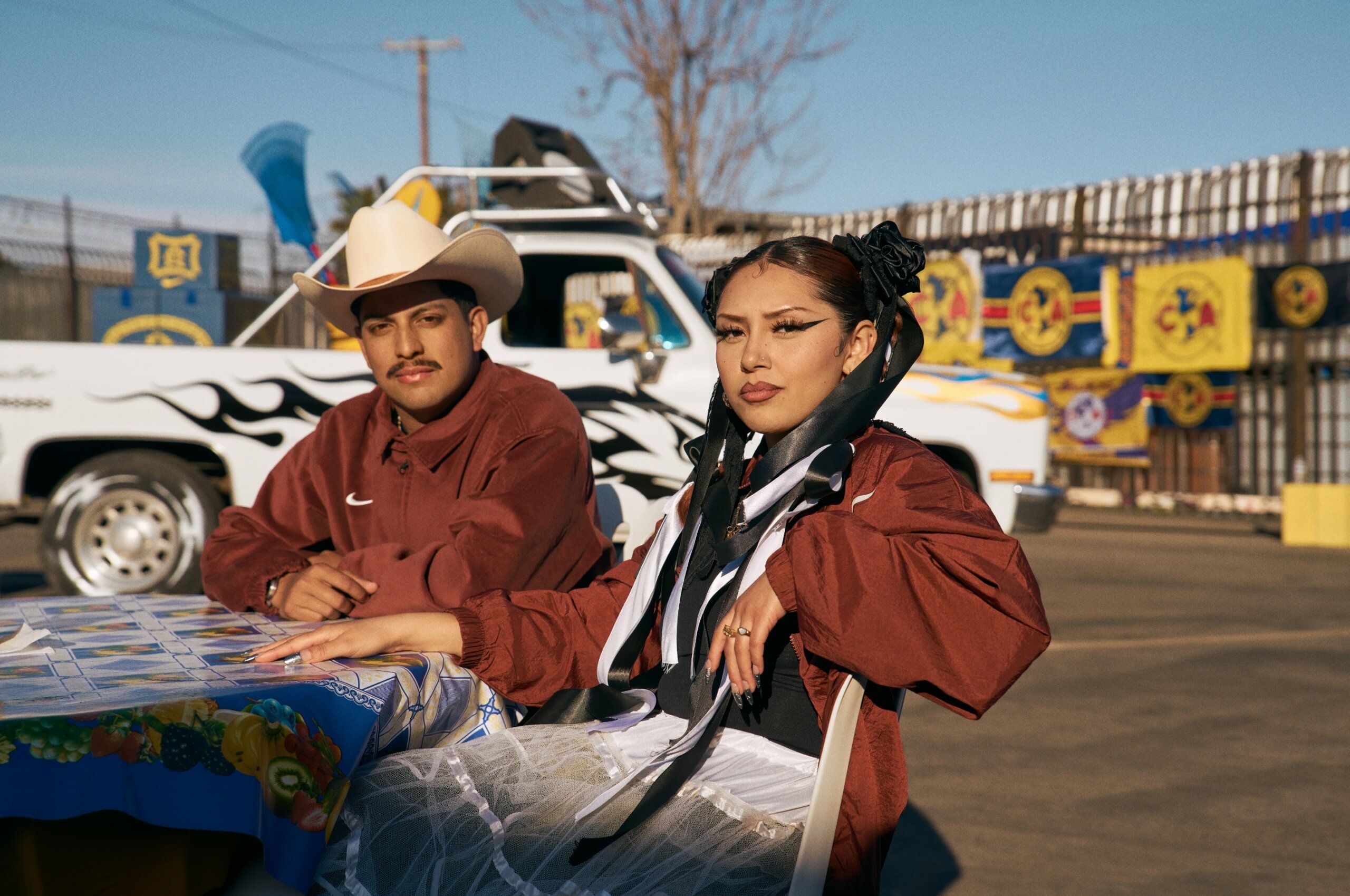 Two people sit at a table outdoors, both wearing maroon jackets. A white pickup truck and colorful banners are visible in the background.