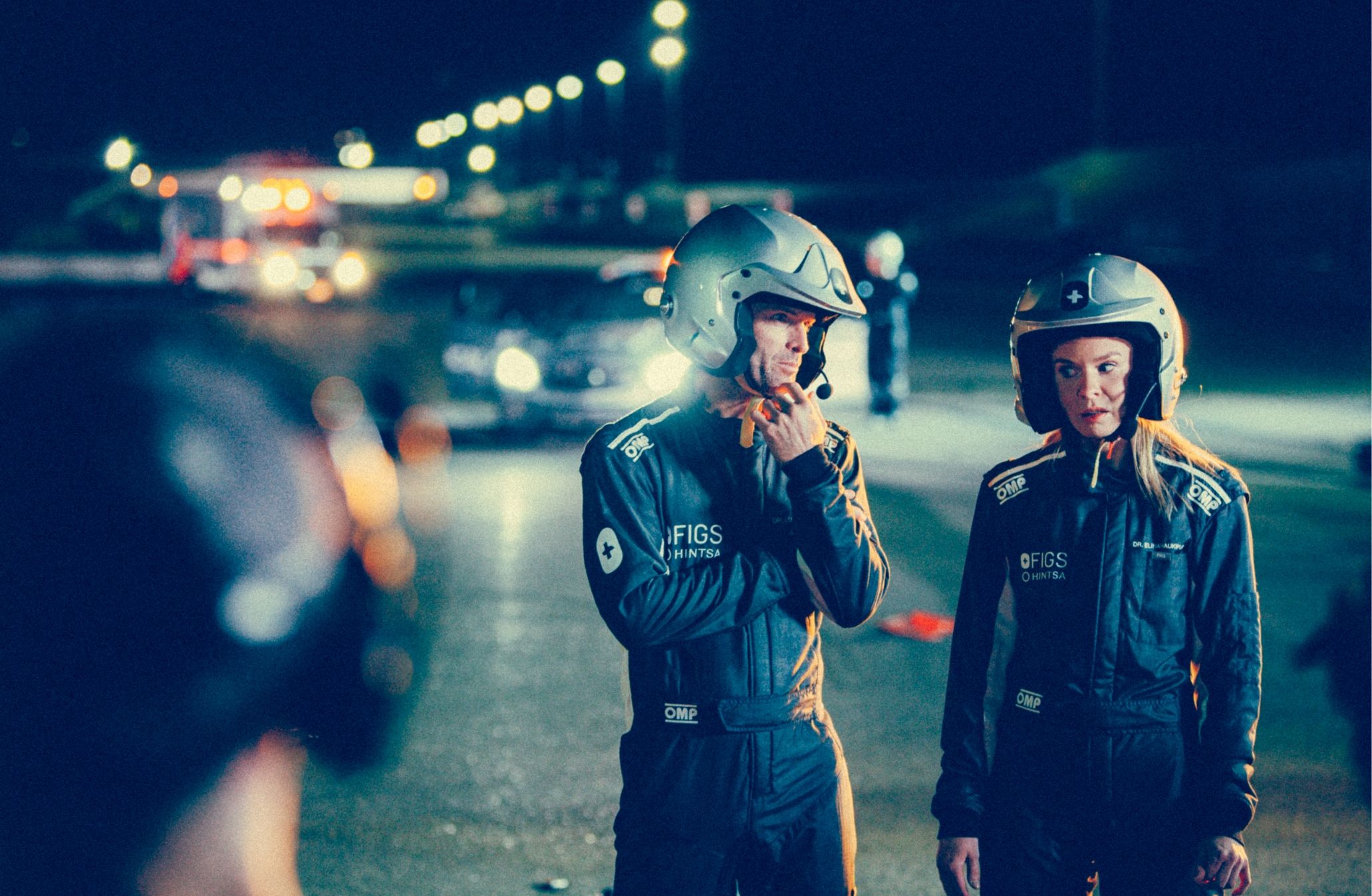 Two race car drivers in helmets and jumpsuits stand together on a lit track at night, with emergency vehicles in the background.