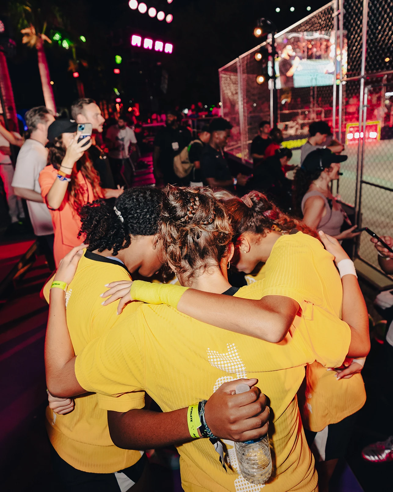 Three people in yellow shirts huddle together near a sports field at a lively, colorful nighttime event with spectators and lights.