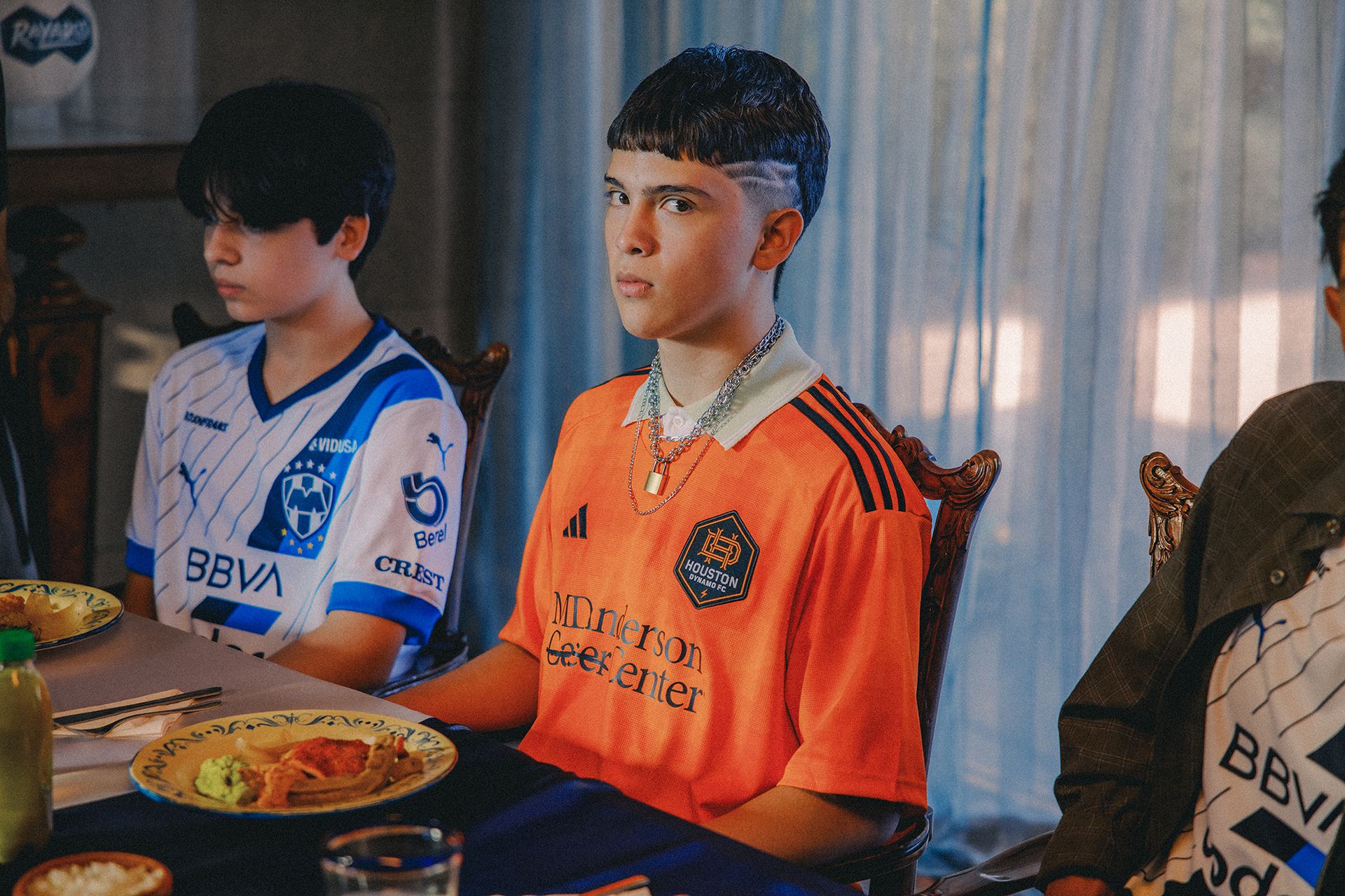 Two young individuals sit at a dining table. One wears an orange soccer jersey, and the other wears a blue and white striped soccer jersey. Plates of food are set in front of them.