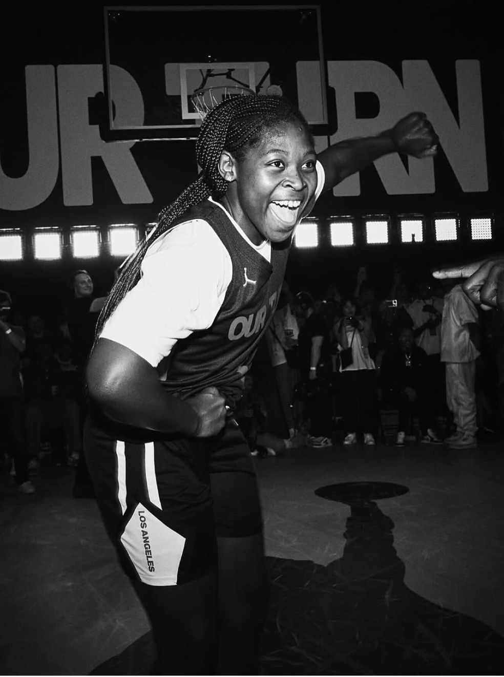 A basketball player in a Los Angeles uniform smiles and poses energetically on a court with a cheering crowd in the background.