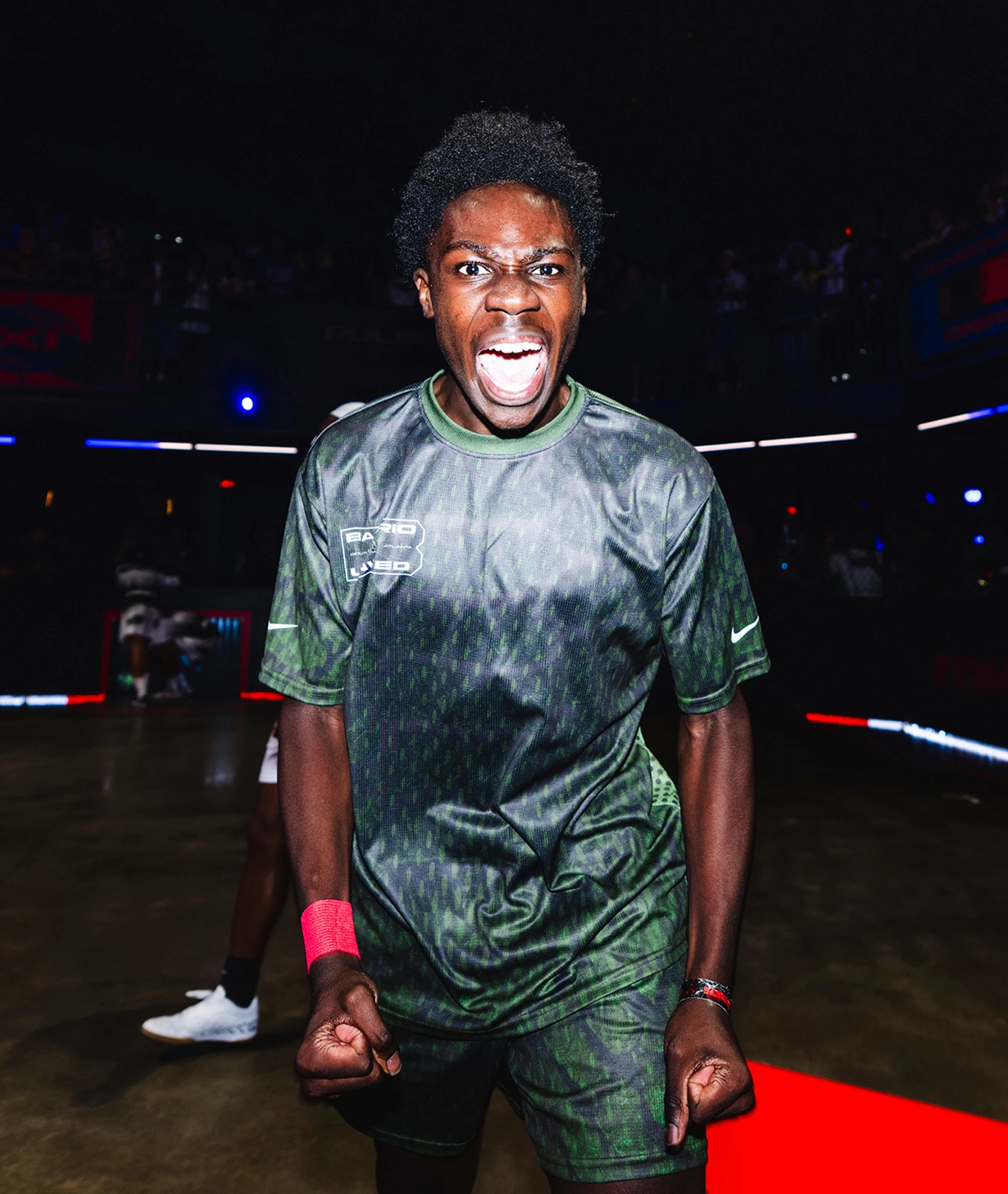 A young athlete in a green uniform clenches his fists and shouts energetically on an indoor court, with dramatic lighting and blurred spectators in the background.