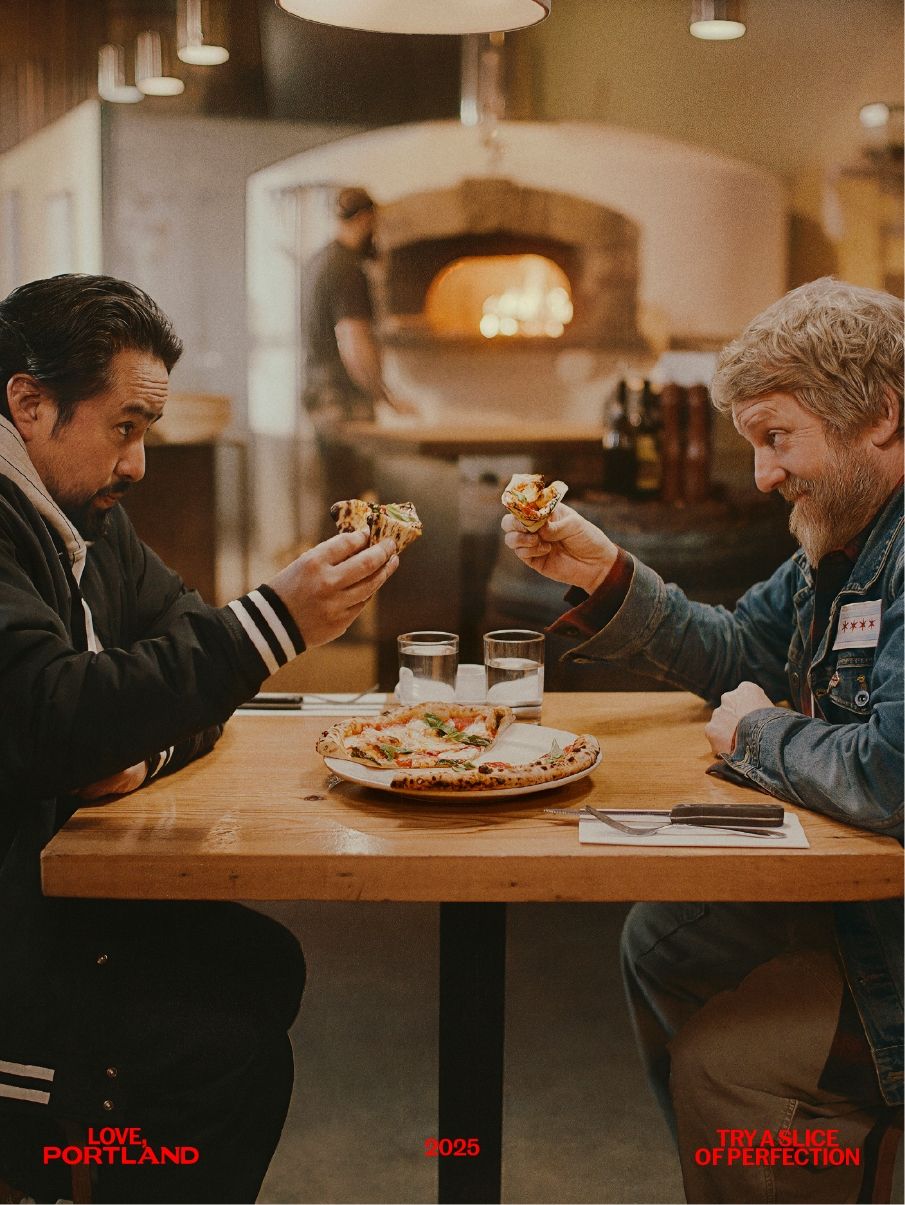 Two men sit across from each other at a table in a pizza restaurant, raising pizza slices in a toast, with a wood-fired oven and a chef in the background.