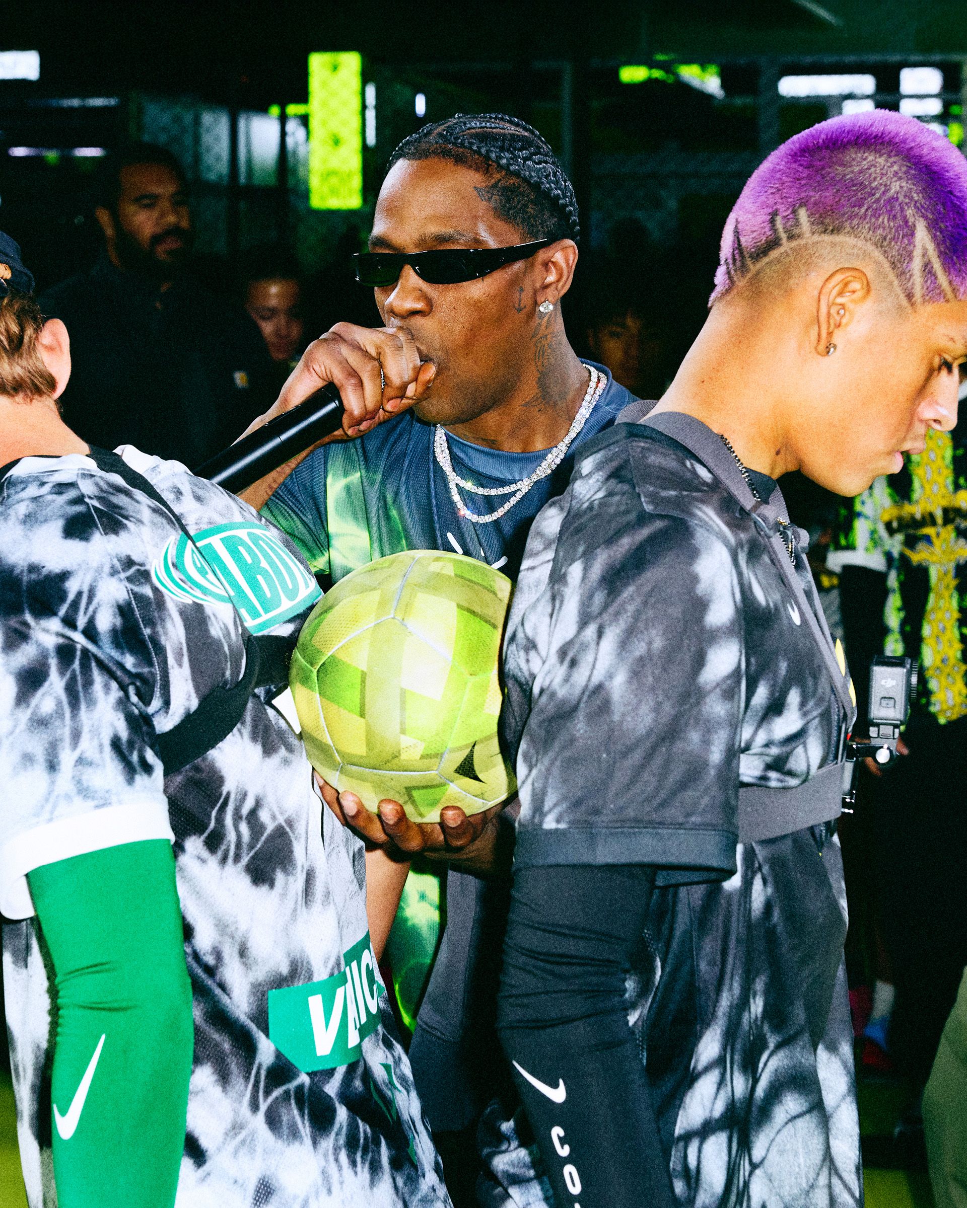A man wearing sunglasses holds a microphone and a green ball, flanked by two people in tie-dye shirts, at an indoor event.