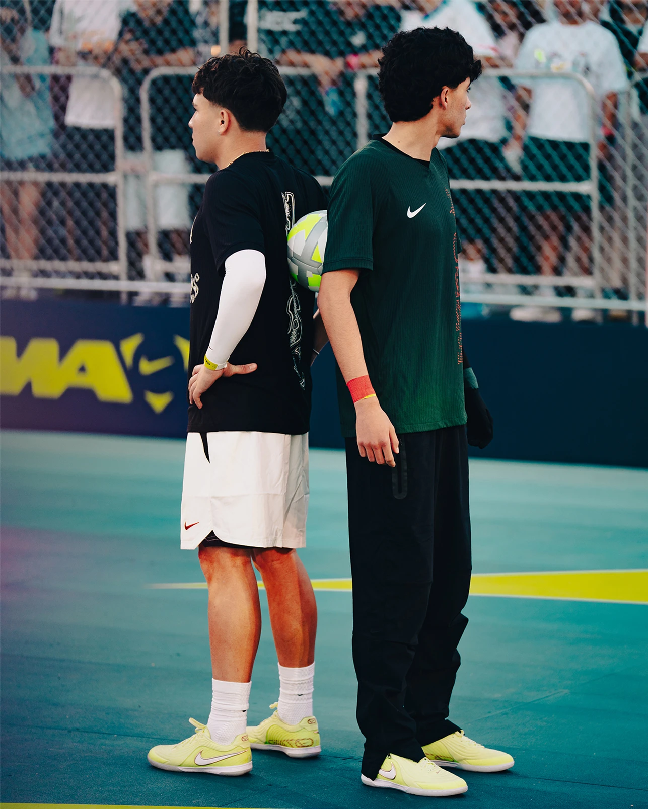 Two young men stand back to back on an outdoor sports court, both wearing athletic clothing and neon shoes, while one holds a soccer ball. A chain-link fence and spectators are in the background.