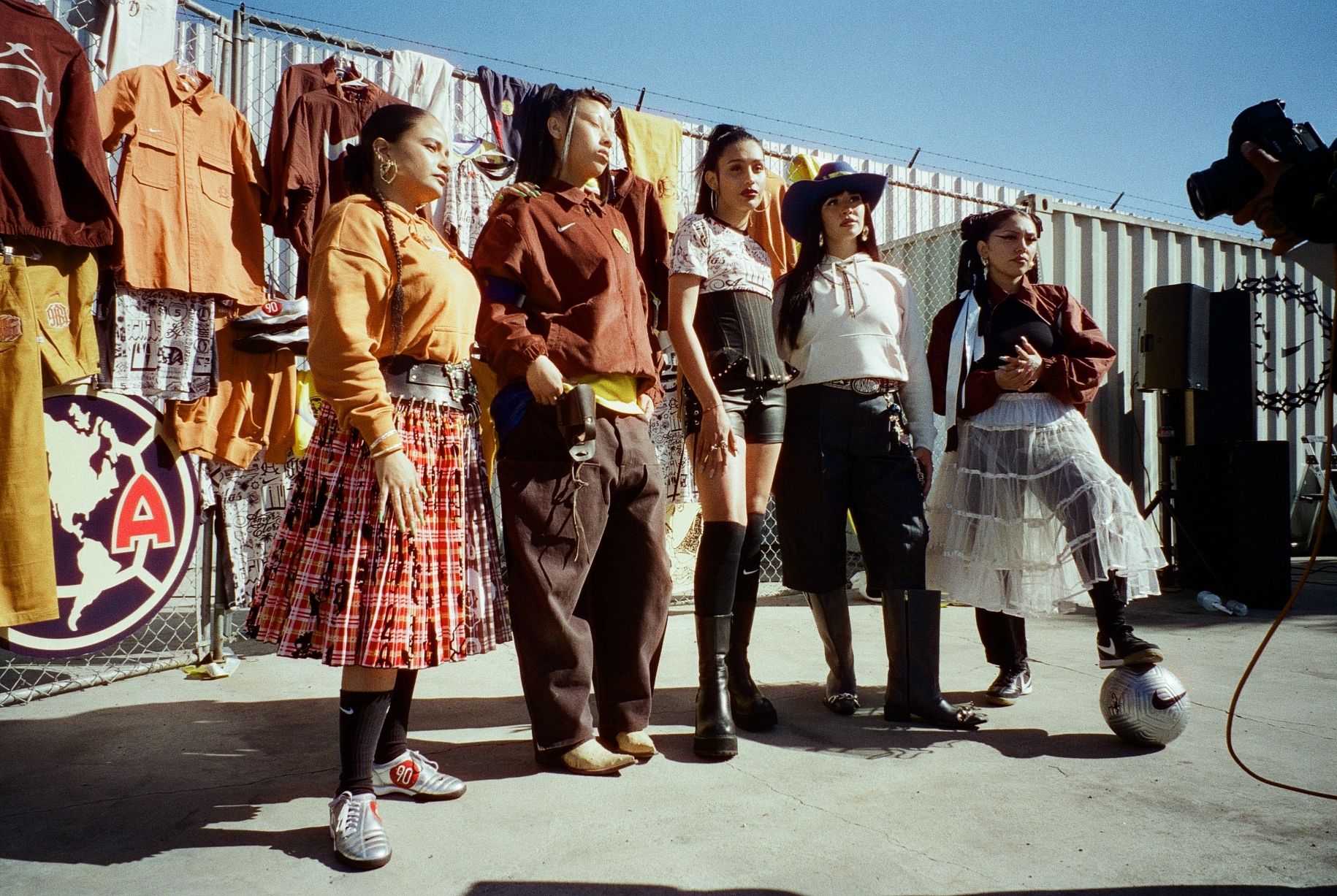 Five people pose together outdoors in front of a fence displaying clothing, with sunlight casting shadows and one person standing near a soccer ball.