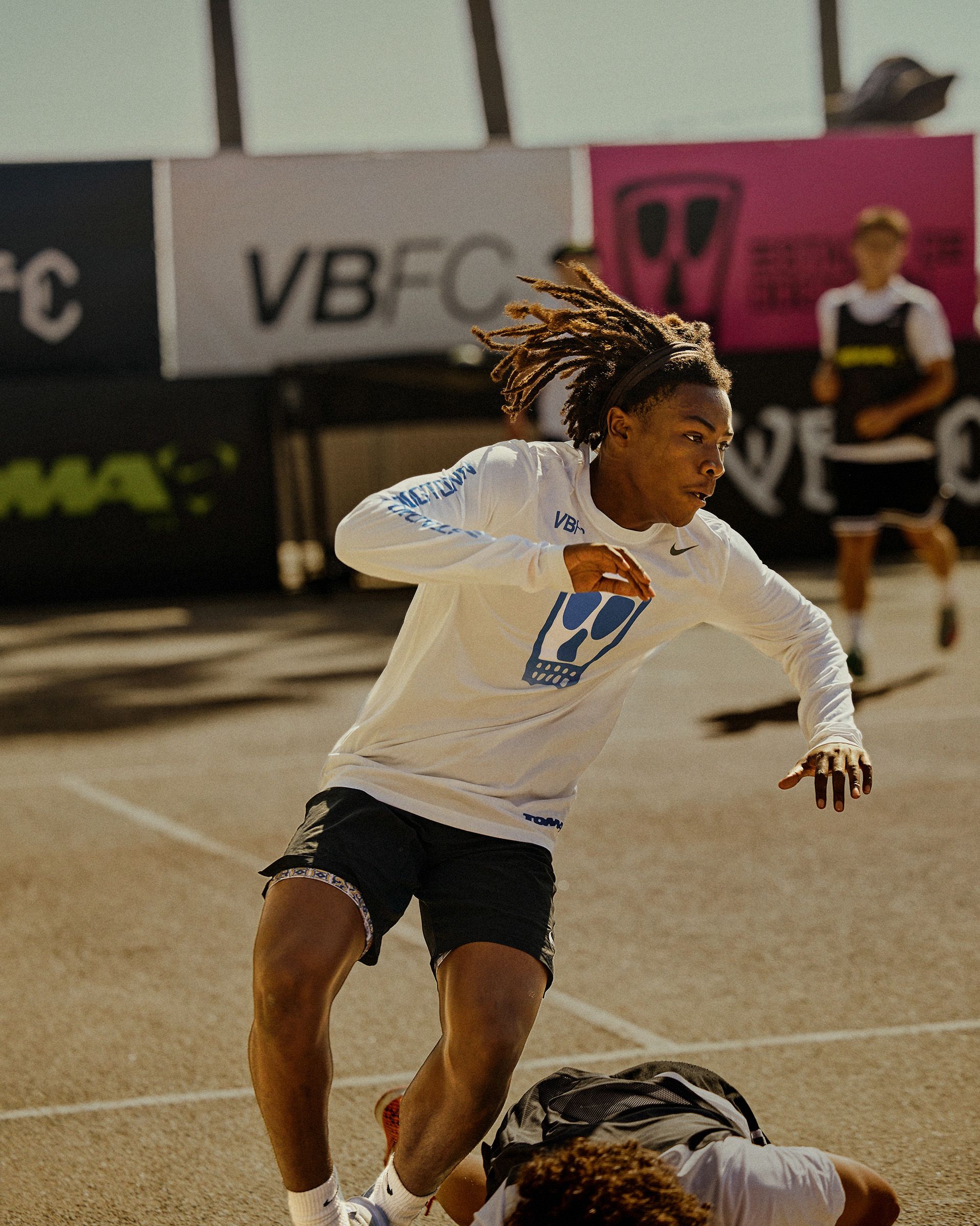 A soccer player in a white long-sleeve shirt and black shorts runs on an outdoor court, as another player falls to the ground in front of him.