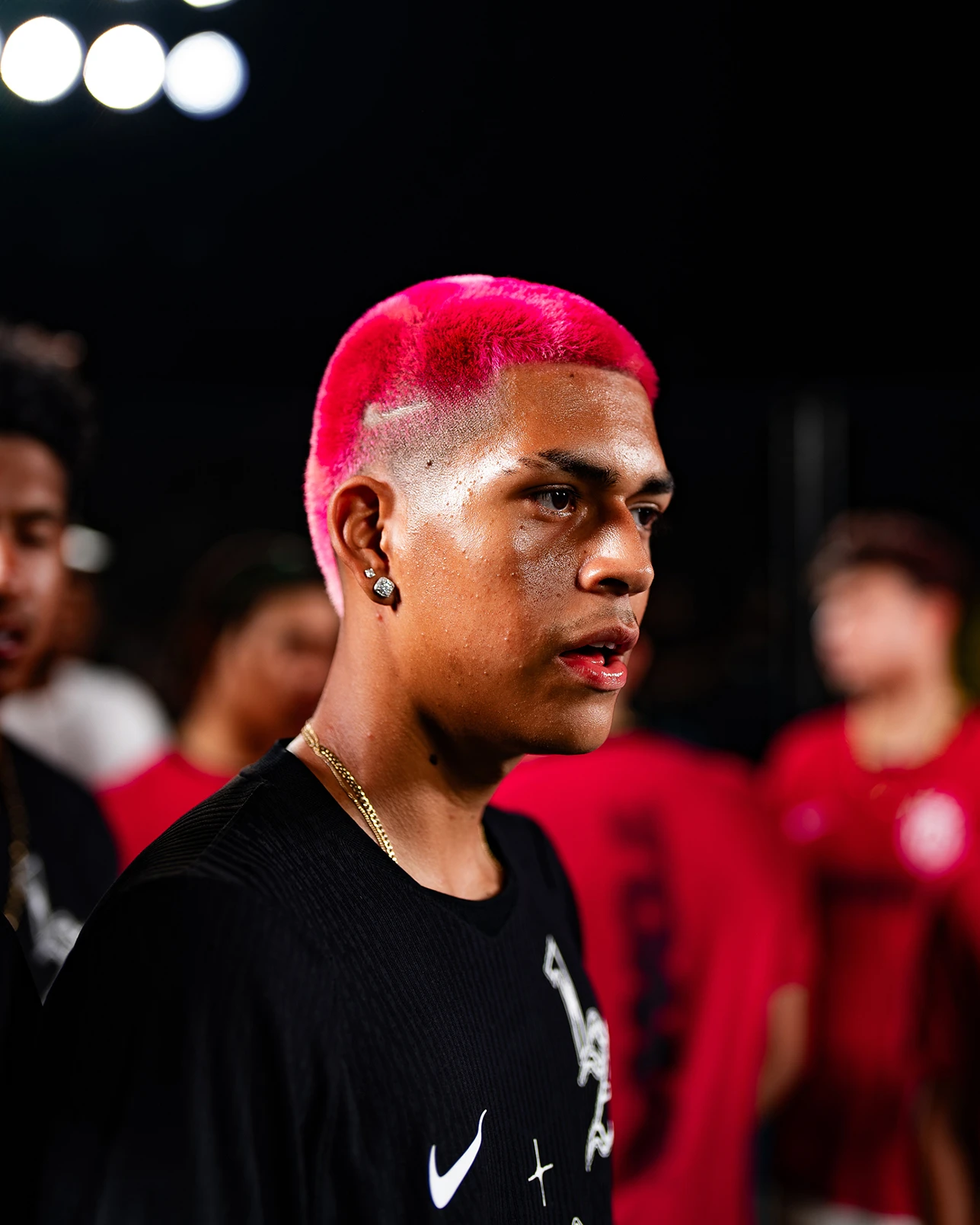 A young man with short, bright pink hair and earrings stands in profile wearing a black Nike shirt, with blurry people and lights in the background.