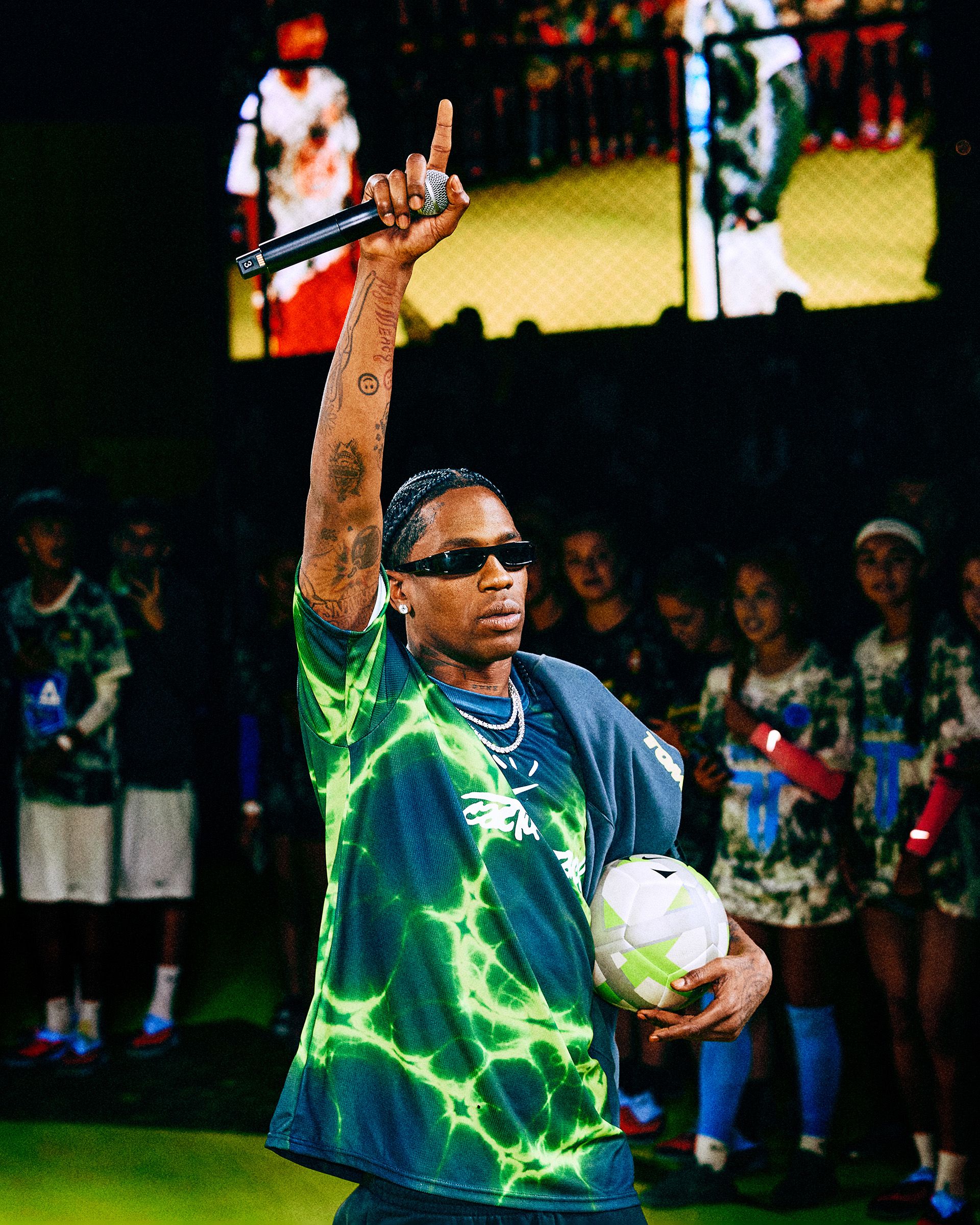 A man wearing sunglasses and a green tie-dye shirt holds a soccer ball in one hand and raises the other hand with a microphone, standing in front of a group of people.