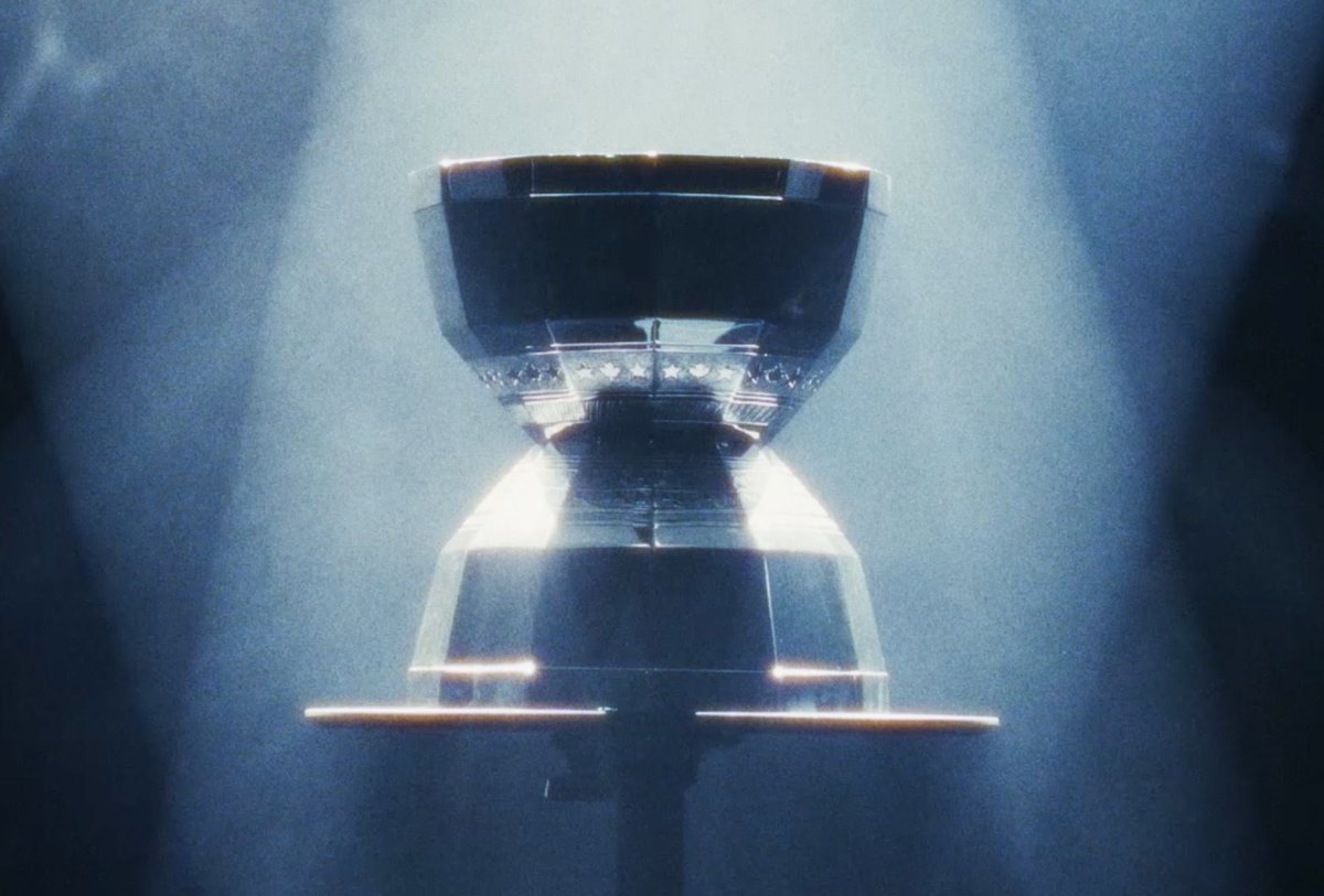 A metallic, hourglass-shaped object is displayed on a stand, illuminated by beams of light in a dark, smoky environment.