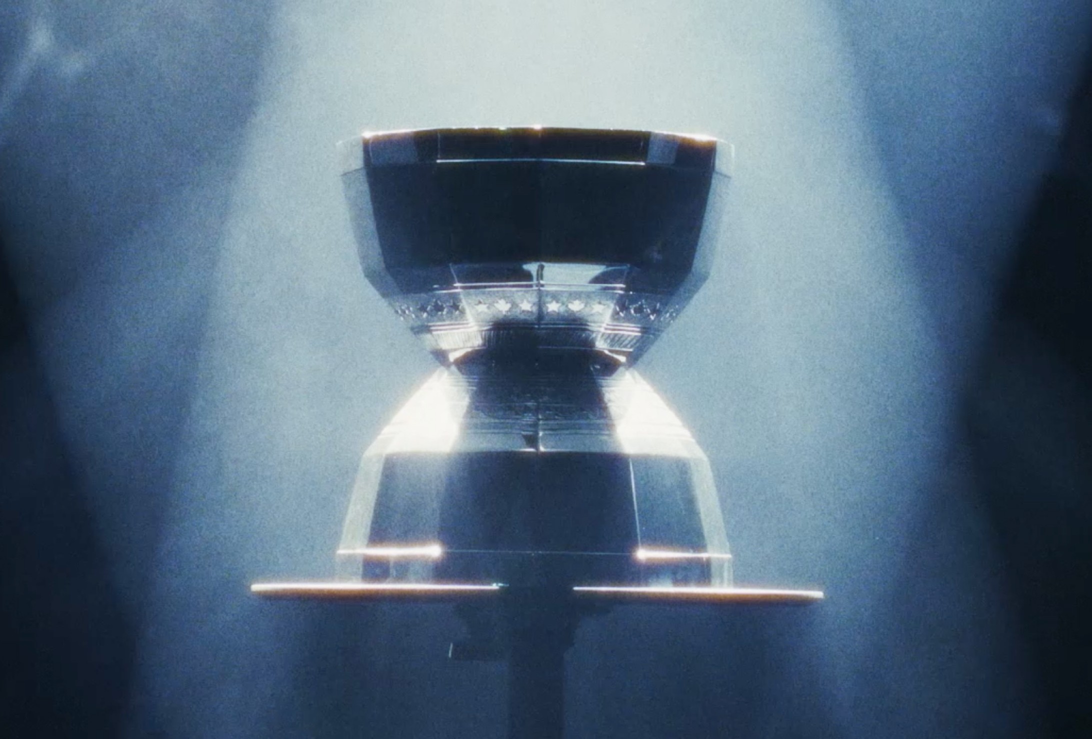 A metallic, hourglass-shaped object is displayed on a stand, illuminated by beams of light in a dark, smoky environment.